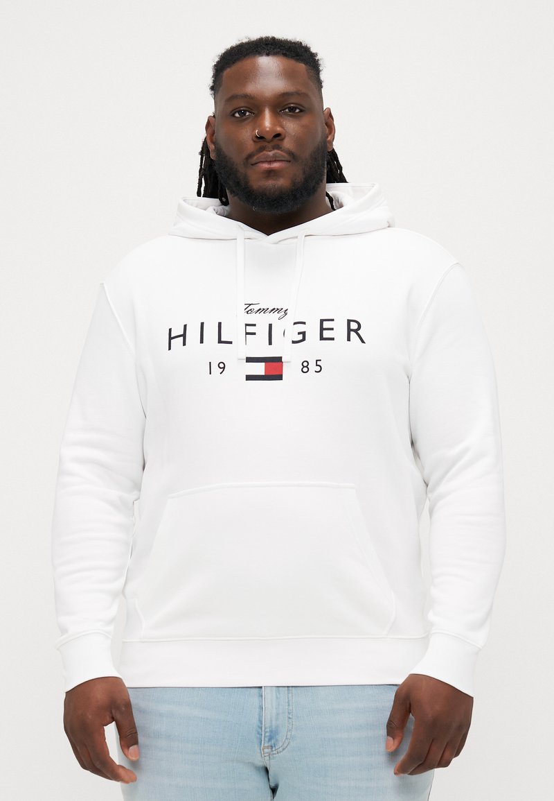 Biela mikina s predným vreckom a sťahovacou kapucňou. Obsahuje čierny nápis "Tommy HILFIGER" a logo v červenej, modrej a bielej farbe. Mäkká textúra látky.