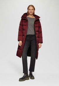 Burgunderfarbene gepolsterte Jacke mit Kapuze, graues Langarmshirt, schwarze Jeans und schwarze Stiefel mit dicker Sohle, ausgestattet mit einem durchgehenden Reißverschluss und Knöpfen.