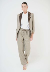Donna che cammina con blazer beige a quadri dal colletto marrone, pantaloni a gamba larga coordinati, camicia bianca e tacchi aperti dorati su sfondo bianco.