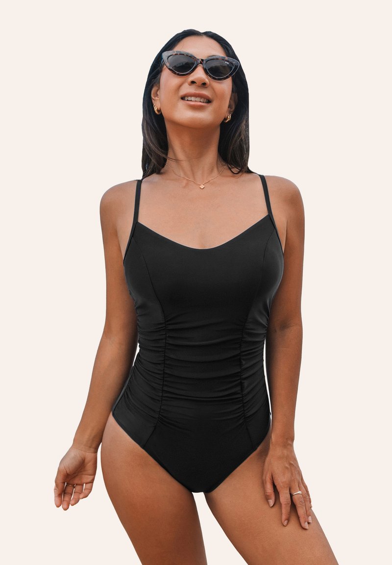 Maillot de bain noir une pièce avec bretelles réglables, détails froncés, encolure en V classique et texture lisse. Porté avec des lunettes de soleil en forme de chat.