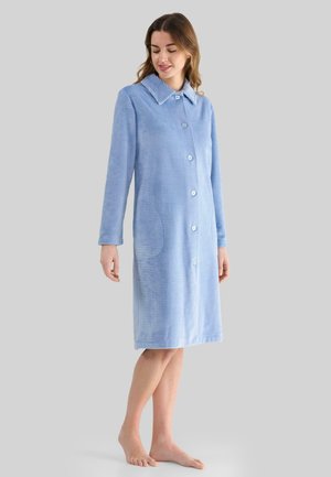 Mujer vestida con una bata de botón de textura ligera de color azul claro, de longitud hasta la rodilla, con cuello y bolsillos laterales, de pie descalza sobre un fondo liso.