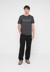 Γκρι βαμβακερό μπλουζάκι με πράσινο σχέδιο "Timberland Established 1973", συνδυασμένο με μαύρο παντελόνι και μπεζ μποτάκια αστραγάλου. Απλός και καθημερινός σχεδιασμός.