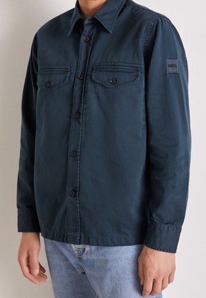 Man draagt een marineblauw overhemd met knopen, met twee zakken op de borst met knopen en een BOSS-logo patch op de linkermouw, gecombineerd met lichtblauwe jeans.