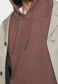 Brauner Hoodie mit Textur, Kordelzug und braunen Enden, darunter eine beigefarbene Jacke mit Knopf und schmalem Revers getragen.