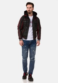 Schwarze und burgunderrote Kunstlederjacke mit Kapuze, mehreren Taschen und roten Akzentstreifen; kombiniert mit verwaschenen blauen Jeans und schwarzen Sneakers.