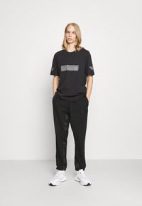 Sort bomulds t-shirt med en tekstureret Adidas-logo på brystet, parret med sorte joggers med slanke sidestriber og hvide sneakers.