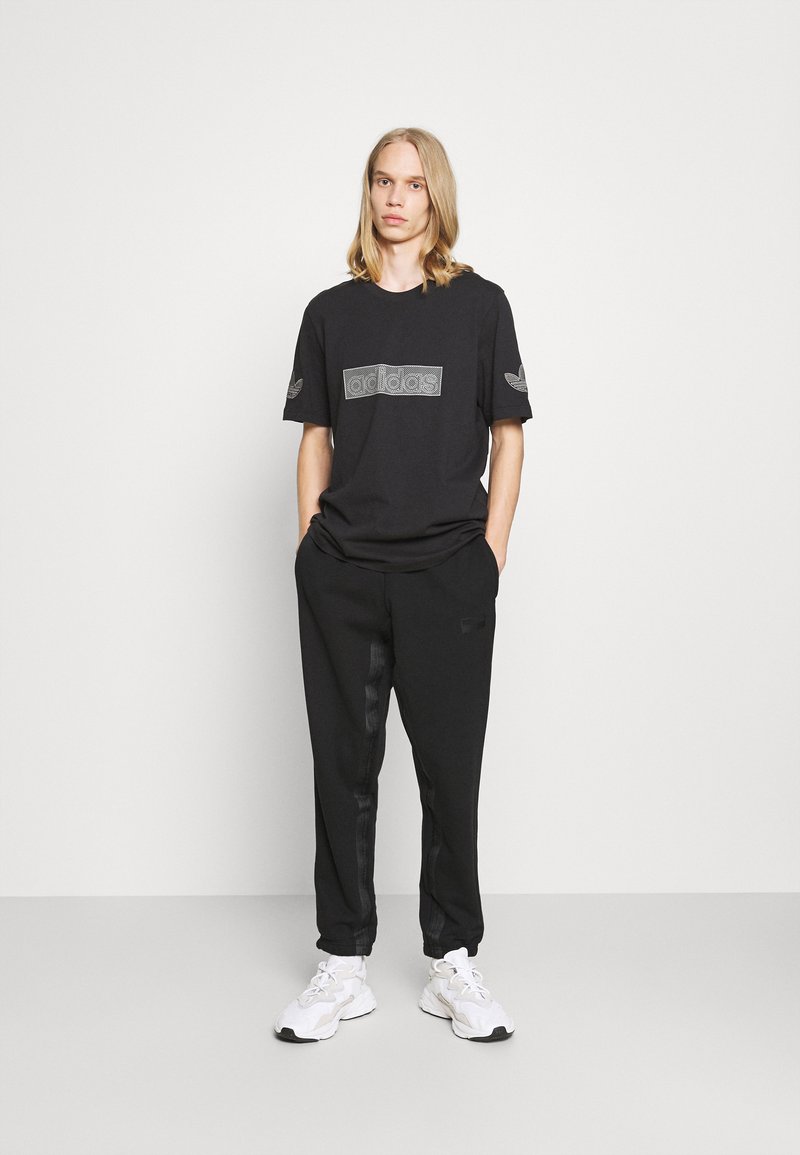 Sort bomulds t-shirt med en tekstureret Adidas-logo på brystet, parret med sorte joggers med slanke sidestriber og hvide sneakers.