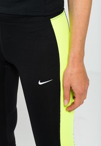 Legginsy Nike w czarnym kolorze, z jasną neonową żółtą boczną wstawką, wykonane z elastycznego materiału, z akcentem w postaci białego logo.