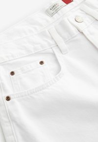 Pantalon en coton blanc avec une taille mi-haute, présentant une poche avant et des rivets en bronze décoratifs. Le tissu a une texture subtile.