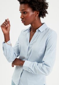 Chemise boutonnée rayée bleu clair avec col, manches longues avec poignets froncés et une poche poitrine. Texture de tissu lisse.