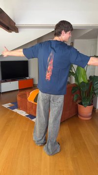 Mann mit kurzen braunen Haaren, der ein dunkelblaues Hemd mit einem roten Flammendesign auf dem Rücken trägt und mit ausgebreiteten Armen in einem Wohnzimmer steht.