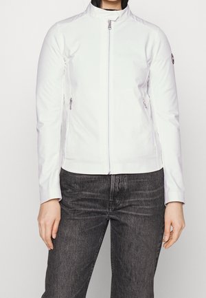 Donna che indossa una giacca bianca con cerniera e collo alto, e jeans neri, in piedi davanti a uno sfondo semplice.