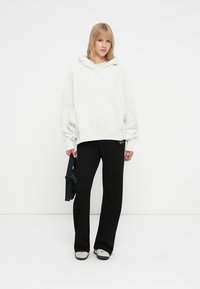 Hoodie oversized branco com um acabamento texturizado, combinado com calças pretas com nervuras e sapatilhas cinzentas. Modelo segura uma bolsa preta. Look simples e moderno.