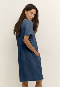 Blue chambray dress with short sleeves, relaxed fit, side pockets, and pleated detail at the waist. Soft, textured fabric.