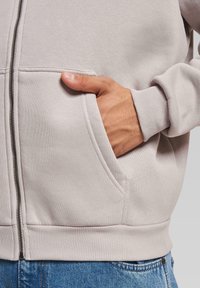 Lys grå hettejakke med glidelås laget av mykt stoff. Den har ribbede mansjetter, en frontlomme og synlige sømdetaljer. Bæreres over blå jeans.