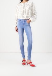 Ljusblå skinny jeans med hög midja, i kombination med en krämfärgad blus med rynkor och röda högklackade mules med spetsiga tår och detaljer.