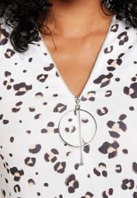 Vit plagg med leopardtryck, utrustat med en lång silvrig dragkedja och cirkelformad dragkedjehandtag. Slät textur, design med v-ringning.