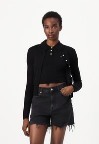 Svart ribbestrikket cardigan med krage, knappet foran. Brukt med en matchende svart kort topp og slitte svarte denimshorts.