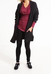 Langer schwarzer Cardigan, burgunderfarbenes Oberteil mit Drapée-Ausschnitt und Knopfakzenten, schwarze Leggings und schwarze Sportschuhe. Die Textur wirkt weich.