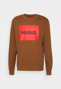 Sweat-shirt marron avec un grand graphique rectangulaire rouge portant le mot "HUGO" en noir. Col rond et poignets côtelés.
