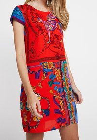 Czerwona sukienka z krótkim rękawem, zdobiona żywymi wzorami, w tym motywem wielbłąda i kwiatów, o zwiewnym kroju i różnorodnych kolorowych akcentach.