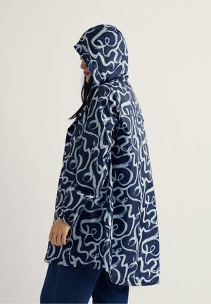 Žena u tamnoplavoj kišnoj jakni s apstraktnim bijelim valovitim uzorkom i kapuljačom, stoji u profilu na jednobojnoj pozadini.