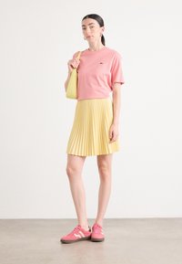 T-shirt di cotone rosa, gonna gialla plissettata e sneakers rosa con accenti bianchi. Borsa a tracolla gialla. Sfondo neutro.