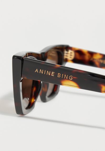 Κοντινό πλάνο γυαλιών ηλίου με καφέ μάρμαρο και μαύρα περιγράμματα, με χρυσά γράμματα "ANINE BING" στο πλευρικό σκέλος.