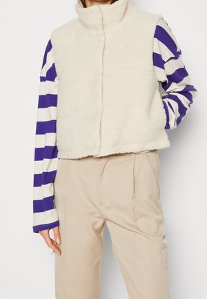 Persoon draagt een crèmekleurig mouwloos fleecevest over een paars-wit gestreept shirt met lange mouwen en beige broek met hoge taille.