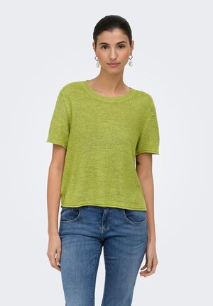 Kvinde med mørkt hår i en grøn strikket top med korte ærmer og blå jeans, stående mod en ensfarvet lys baggrund.
