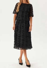 Schwarzes Spitzenkleid mit floralem Muster, kurzen Ärmeln und schichtierter Gestaltung, in knielanger Länge und kombiniert mit schwarzen High Heels.