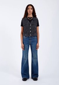 Czarna kamizelka z denimu z guzikami i dwoma przednimi kieszeniami, zestawiona z rozszerzanymi niebieskimi jeansami i czarnymi botkami na platformie. Gładka faktura i dopasowany krój.