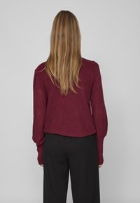 Personne aux longs cheveux châtain clair portant un pull en maille bordeaux et un pantalon noir, debout de dos sur un fond gris uni.