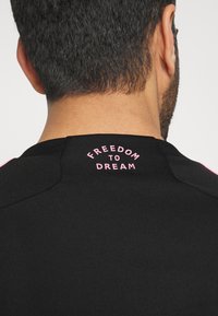 Musta urheilupaita, jossa sileä pinta ja lyhyet hihat, takana niskassa pinkillä painettu teksti "VAPAUS UNELMOIDA".