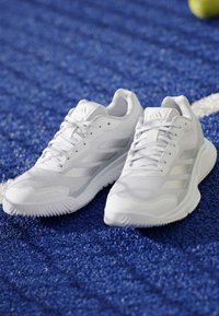Chaussures de sport blanches Adidas avec lacets sur une surface sportive bleue texturée, avec une balle de tennis jaune floue en arrière-plan.