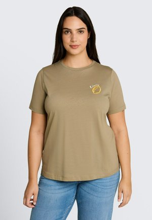 Kobieta o długich ciemnych włosach, ubrana w oliwkowo-zieloną koszulkę z małą żółtą grafiką cytryny i napisem „Limone”, w zestawie z niebieskimi dżinsami.