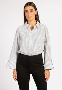 Gestreepte witte blouse met zilveren accenten, ontspannen pasvorm, kraag en klokmouwen. Gecombineerd met zwarte, op maat gemaakte broeken, wat een professionele uitstraling creëert.