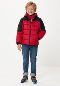 Röd puffjacka med svarta ärmar, dragkedja fram och huva; kombinerad med blå jeans och sneakers. Har en logotyp på vänster axel.