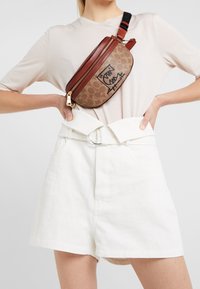 Brun och beige axelväska med logotryck, med dragkedjestängning och en kontrasterande röd detaljer. Justerbar svart rem.