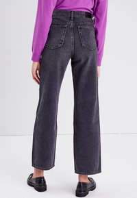 Jeans a vita alta e gamba larga in denim grigio scuro con tasche posteriori e orlo grezzo, abbinati a un maglione viola a maniche lunghe e scarpe nere.