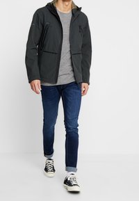 Mørkegrå vandtæt jakke med hætte, to lynlåslommer på brystet og et strømlinet design. Båret over en lysegrå skjorte og blå jeans.