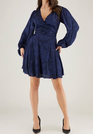 Marineblaue Wickelkleid mit langen Puffärmeln, floralen Textur, tailliertem Schnitt und ausgestelltem Rock, kombiniert mit schwarzen High Heels.
