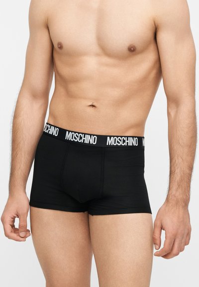 Ανδρικός κορμός με μαύρα boxer Moschino με ελαστική μέση με λογότυπο, στέκεται μπροστά σε απλό ανοιχτό φόντο.