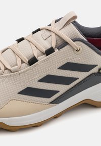 Adidași bej cu partea superioară din țesătură texturată, accente gri închis, design cu dungi negre, sireturi plate și talpă din cauciuc.