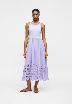 Žena oblečená v bezrukávových levanduľových midi šatách so zvlneným, laserovo rezaným kvetinovým lemom, doplnených hnedými plochými sandálmi a perlovým náhrdelníkom.