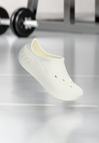 Baltas slip-on apavi ar gludu, teksturētu virsmu, kas aprīkoti ar divām ventilācijas atverēm un izliekta dizaina. Zīmola logo redzams uz sāniem.