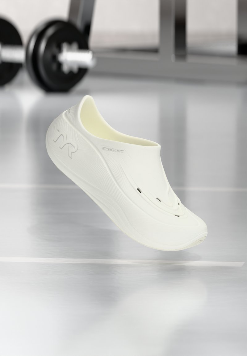Baltas slip-on apavi ar gludu, teksturētu virsmu, kas aprīkoti ar divām ventilācijas atverēm un izliekta dizaina. Zīmola logo redzams uz sāniem.