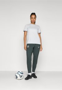 Lysegrå Nike t-skjorte med kort erme, mørkegrønne Nike joggebukser med Chelsea-logo, svarte fotballsko, og en mønstrete fotball på bakken.