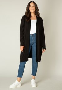 Zwarte cardigan gemaakt van lichtgewicht stof met een open voorkant. Gecombineerd met strakzittende blauwe jeans en witte sneakers, wat een casual stijl toont.
