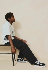 T-shirt graphique blanc avec impression noire, pantalon cargo noir avec poches, baskets noir et blanc, assis sur une chaise noire contre un fond beige.